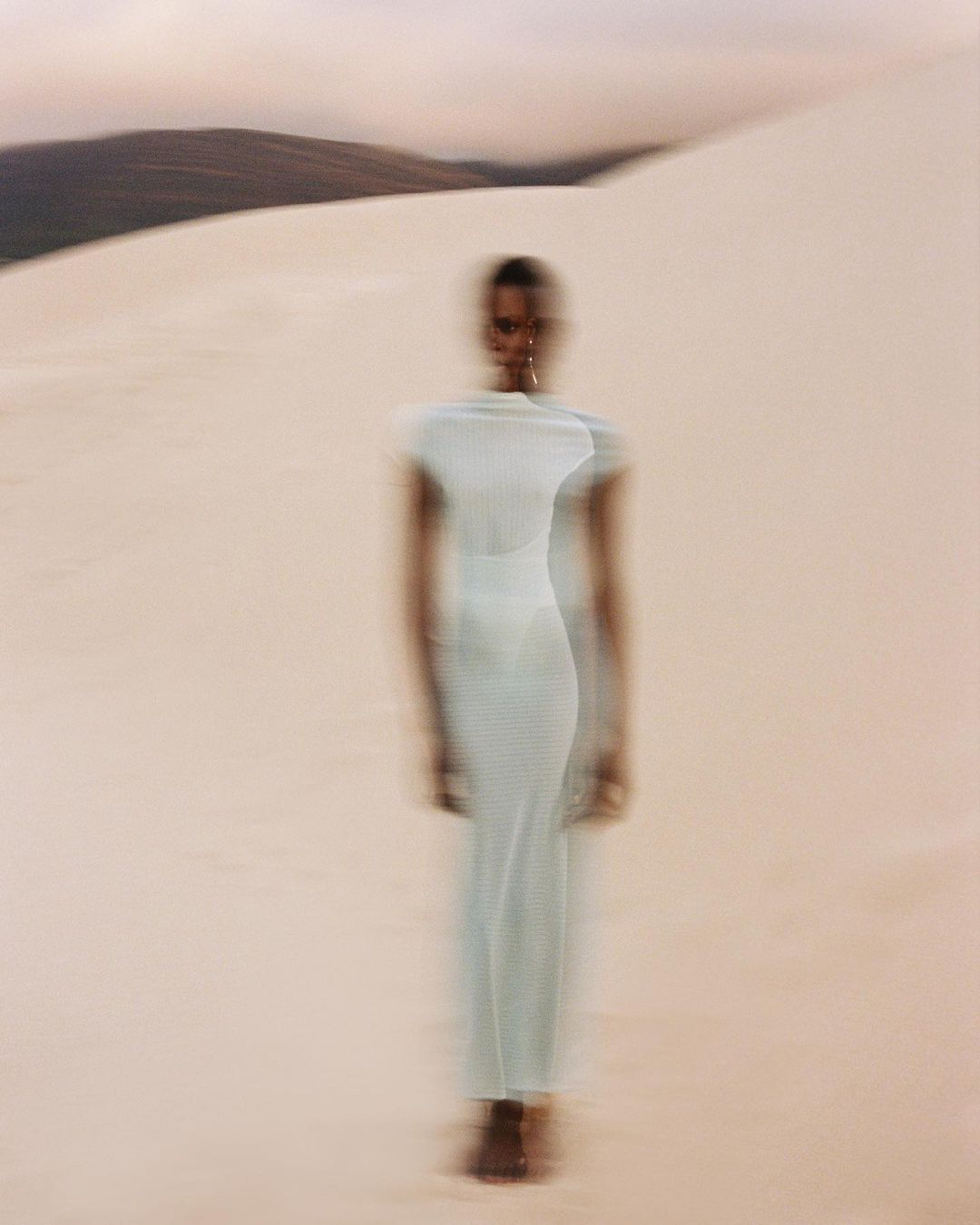Motion-blurred full shot of an African woman in a long dress walking across sand dunes against the background of a mountain ridge.