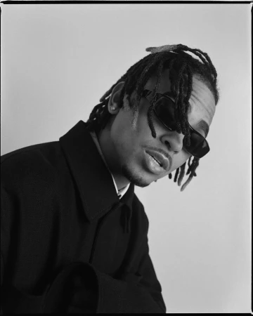 Monochrome portrait of a young Black man with locs wearing sunglasses and a dark overcoat against a neutral backdrop, looking suave.