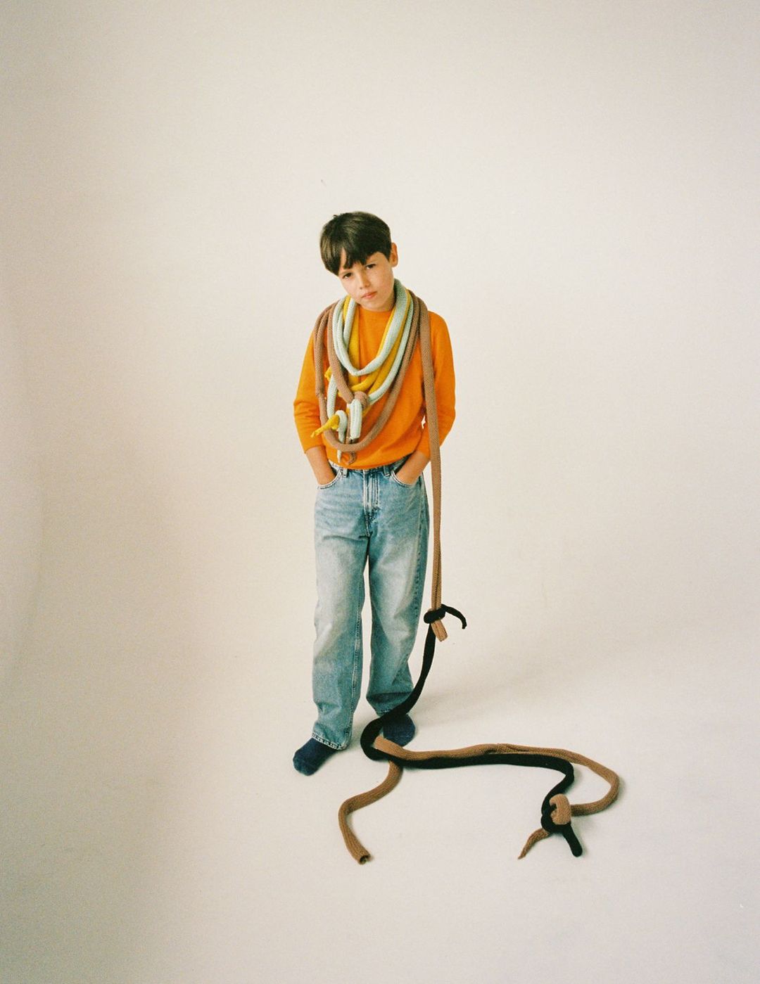 A young person with dark hair standing indoors with a large colorful textile around the neck, wearing an orange shirt and jeans.