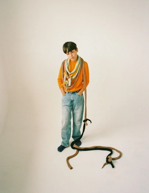 A young person with dark hair standing indoors with a large colorful textile around the neck, wearing an orange shirt and jeans.