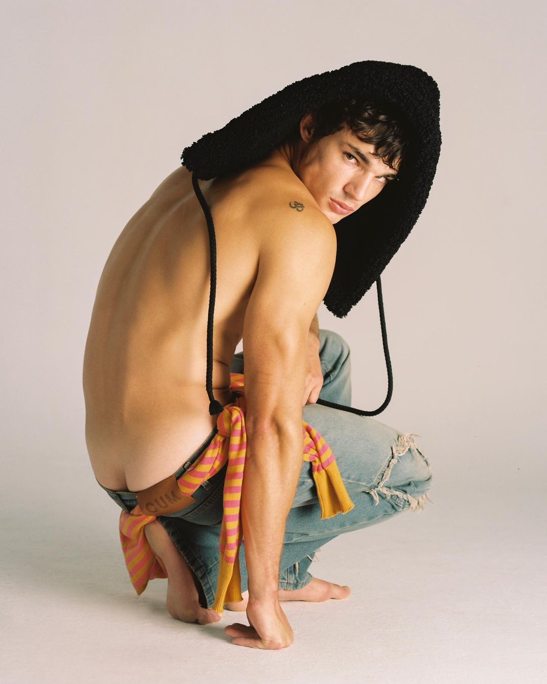 Male model in hooded fashion accessory squats in ripped jeans on a light backdrop in a studio, showing back, shoulder, and bare feet.