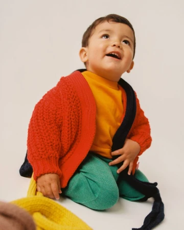Toddler smiles while seated on the floor wearing a colorful knit cardigan, bright yellow t-shirt, and green pants in a studio shot.