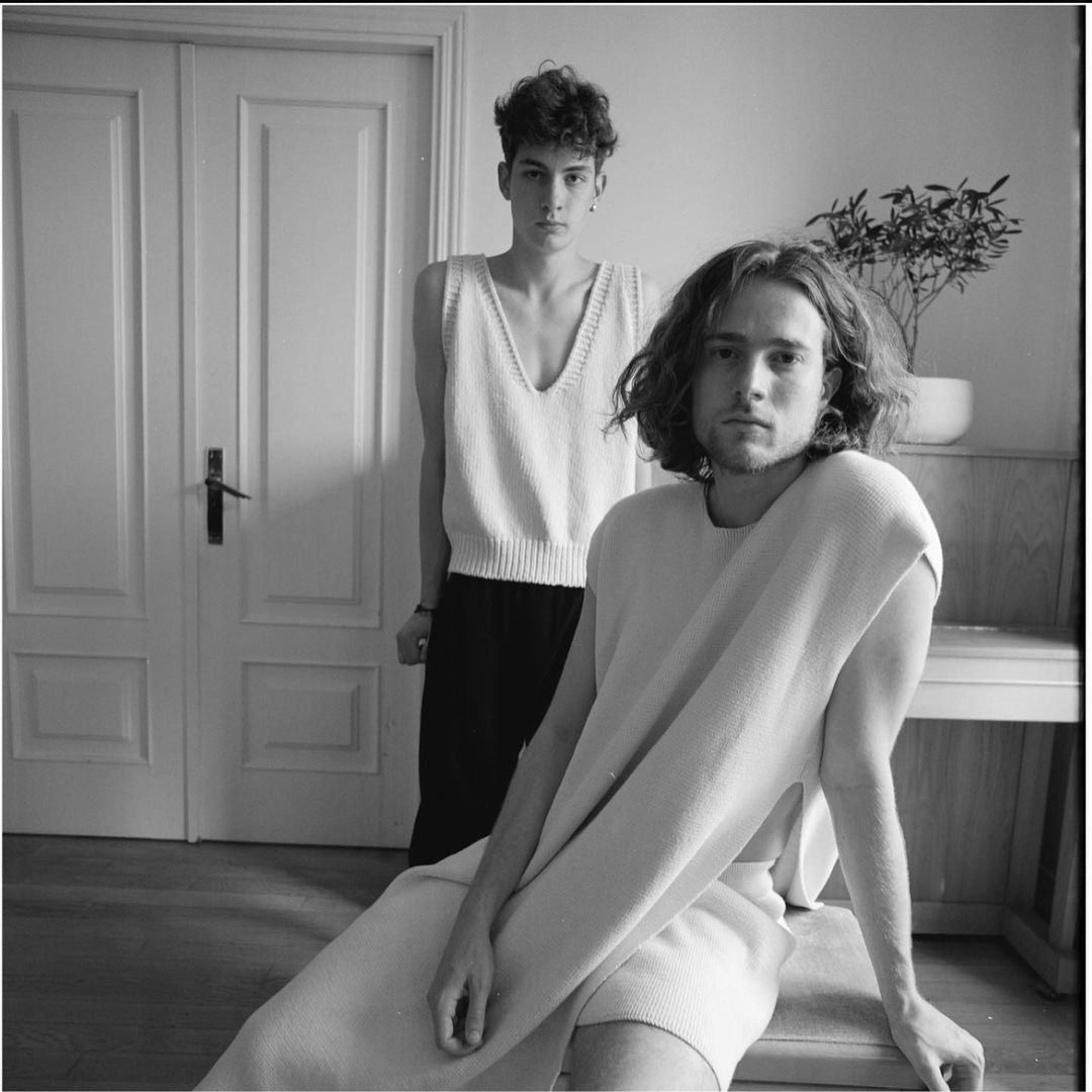 Two fair-skinned young adult models in a black and white studio shot, one seated, the other standing with a plant and door behind.