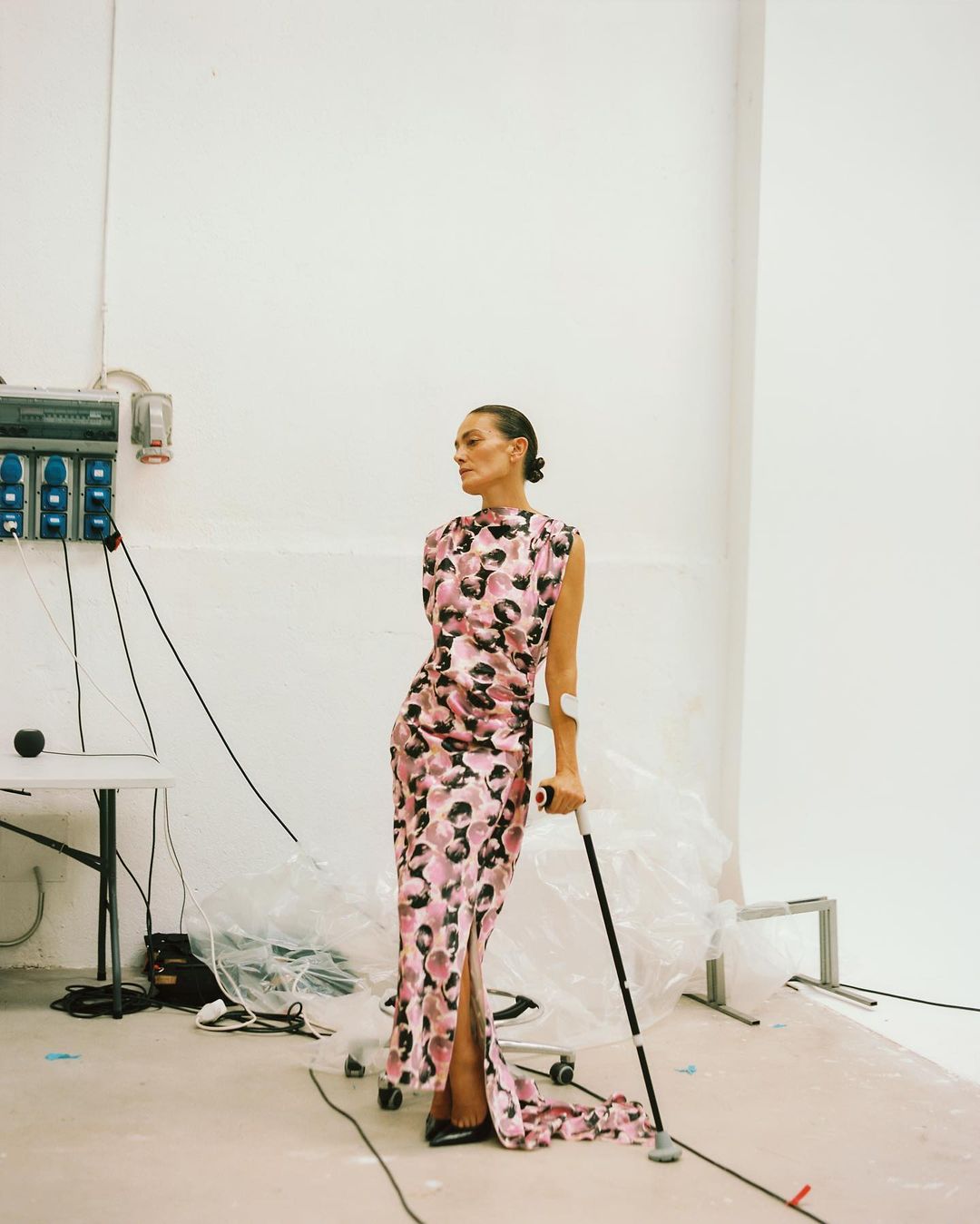 Elegant woman in a patterned gown poses with a crutch in a bright studio. High-fashion meets practicality, capturing a unique portrait.