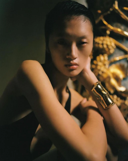 Portrait of a woman with slicked-back hair, wearing a gold cuff bracelet, posing against a textured wall in dim, moody lighting.