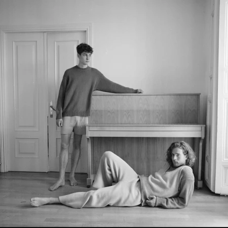 Two young men in an indoor room with a piano, one standing resting an arm on the piano and the other lying on the wooden floor.