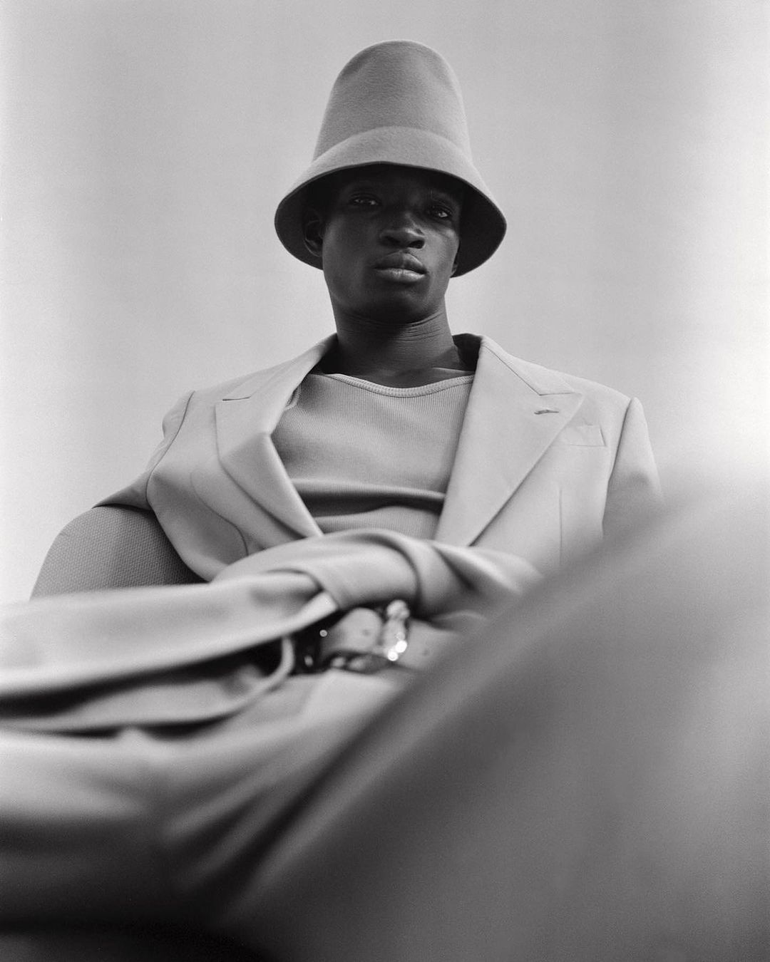 A fashionable Black man in a monochrome beige outfit poses indoors with a funnel-shaped hat and blazer, exuding a calm and sophisticated style.
