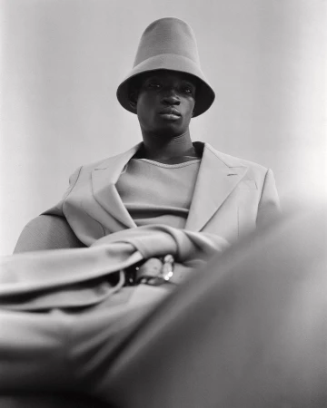 A fashionable Black man in a monochrome beige outfit poses indoors with a funnel-shaped hat and blazer, exuding a calm and sophisticated style.