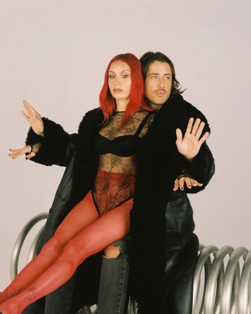 A stylish couple poses together indoors in a light studio setting. The woman with red hair sits in a black fur coat, on a metal object.