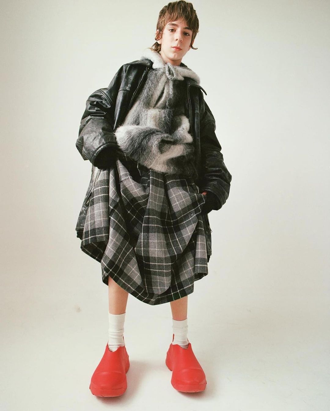Full-length portrait of a child wearing a black leather jacket, grey plaid skirt, gray furry top, white socks, and red shoes against a plain white background.