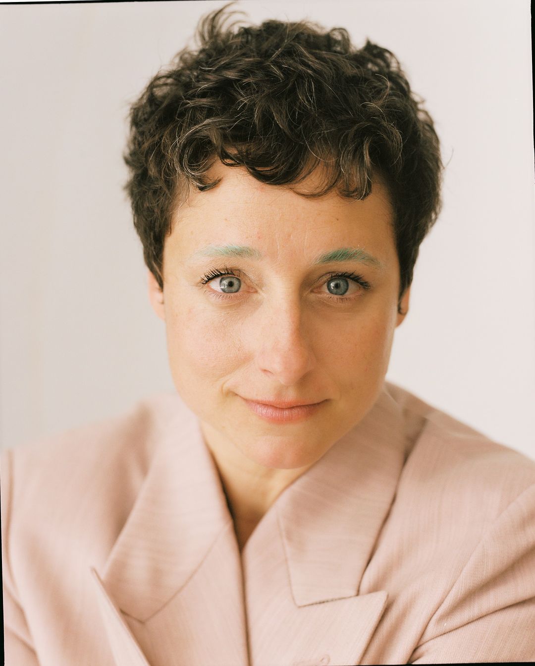Close-up portrait of an adult female with short curly hair and a light pink blazer smiling softly against a neutral background.