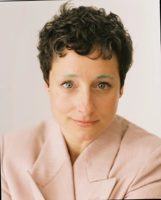 Close-up portrait of an adult female with short curly hair and a light pink blazer smiling softly against a neutral background.