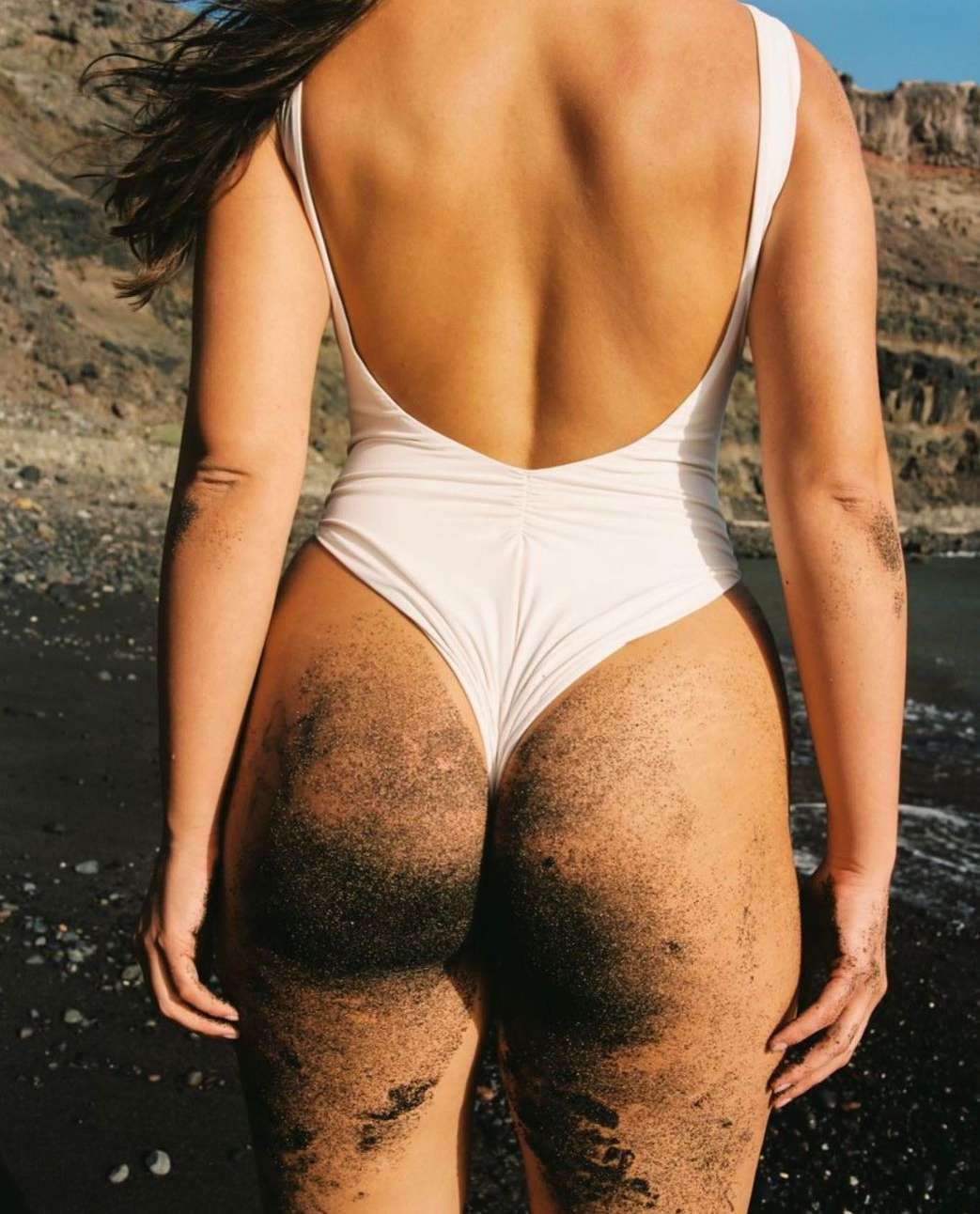 Back view of an adult female in a one-piece swimsuit covered in sand, standing on a black sand beach with a rocky headland in the distance.