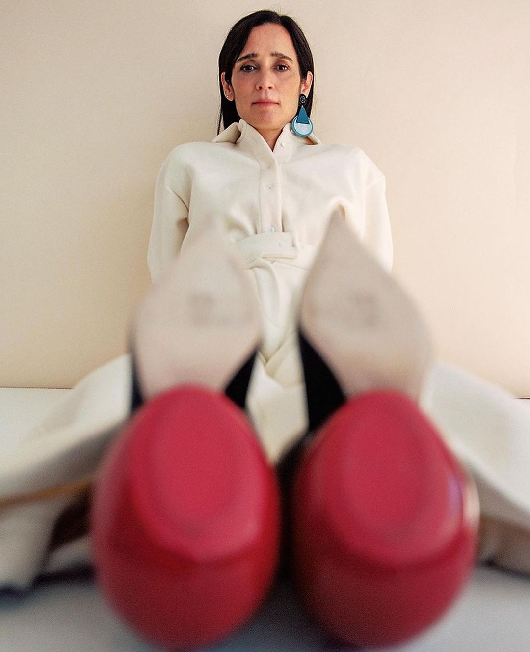 Portrait of a woman in a white jumpsuit and bright red high heel shoes, posing against a light beige background in a photo studio.