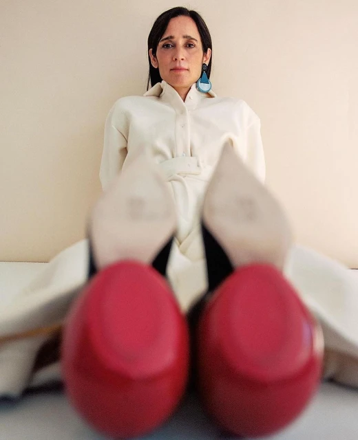 Portrait of a woman in a white jumpsuit and bright red high heel shoes, posing against a light beige background in a photo studio.