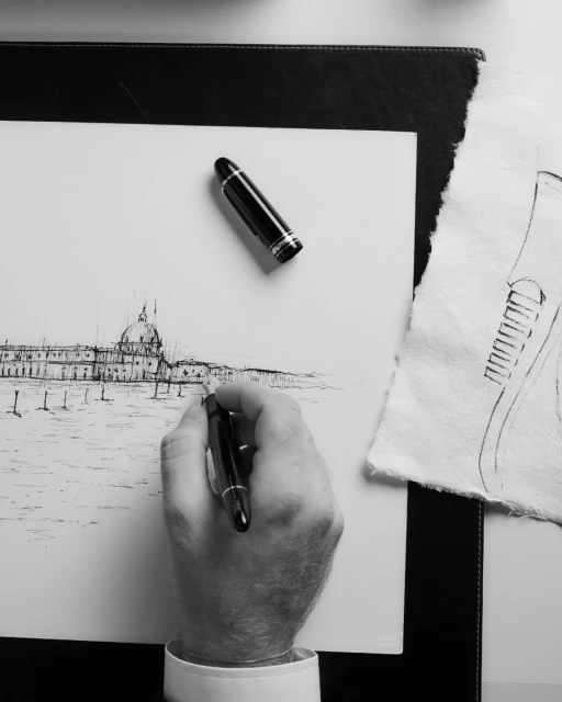Hand drawing a waterscape architecture with a pen on white paper, complemented by a fountain pen and textured sketch, on a dark surface.
