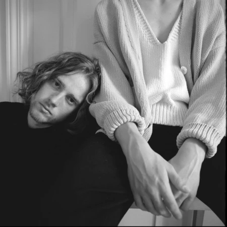Monochrome portrait of a man with curly hair resting his head next to a woman in a knitted sweater with clasped hands indoors.