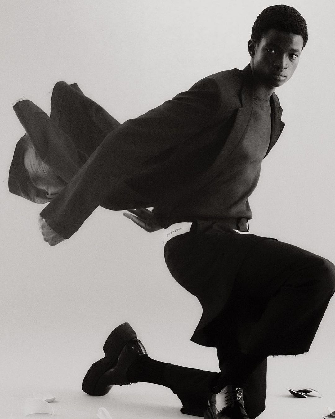 Fashionable Black man posing on one knee in an indoor setting, wearing a dark blazer, sweater, wide-leg pants, and chunky shoes in a monochrome setting.