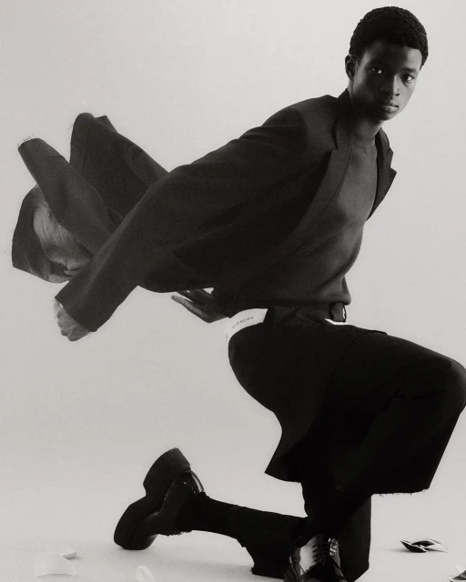Fashionable Black man posing on one knee in an indoor setting, wearing a dark blazer, sweater, wide-leg pants, and chunky shoes in a monochrome setting.