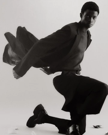 Fashionable Black man posing on one knee in an indoor setting, wearing a dark blazer, sweater, wide-leg pants, and chunky shoes in a monochrome setting.