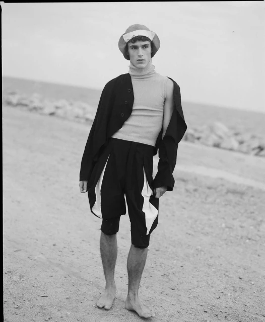 Monochrome portrait of a young adult male fashion model in an unusual hat and outfit posing on a beach with sand and ocean.