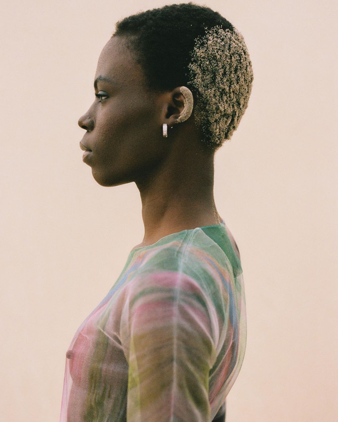 Profile of a young Black woman with dyed blonde hair wearing a semi-sheer, colorful top against a neutral background in a studio setting.