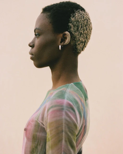 Profile of a young Black woman with dyed blonde hair wearing a semi-sheer, colorful top against a neutral background in a studio setting.