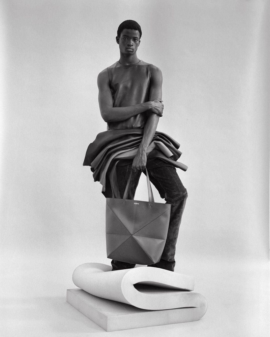 Black-and-white fashion portrait of a Black male model wearing a leather top and jeans, holding a tote bag, studio setting.