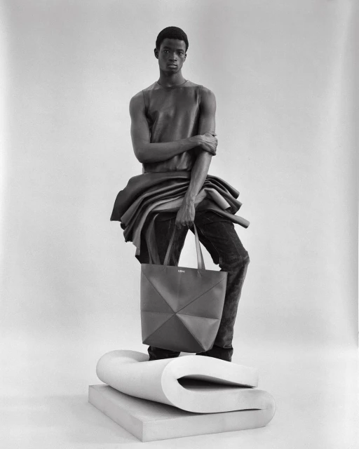 Black-and-white fashion portrait of a Black male model wearing a leather top and jeans, holding a tote bag, studio setting.