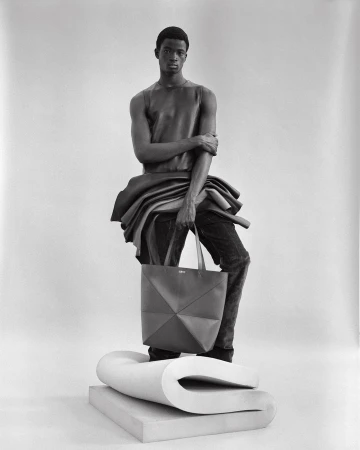 Black-and-white fashion portrait of a Black male model wearing a leather top and jeans, holding a tote bag, studio setting.