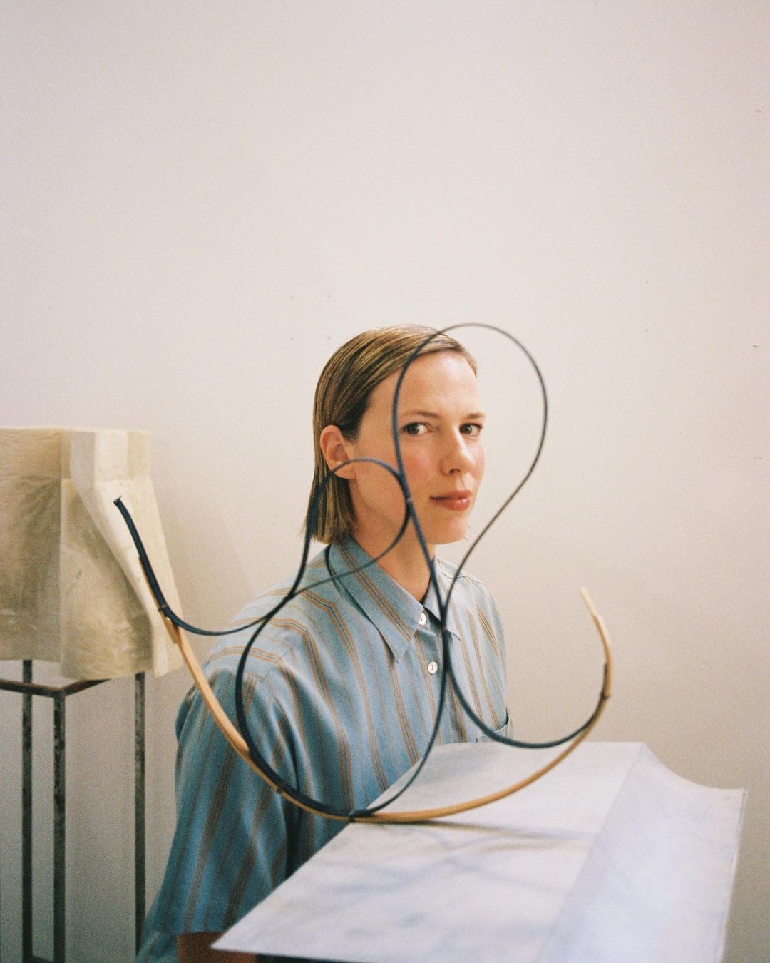 Adult female with short hair poses indoors in front of an abstract sculpture, wearing a striped shirt in a medium studio shot.