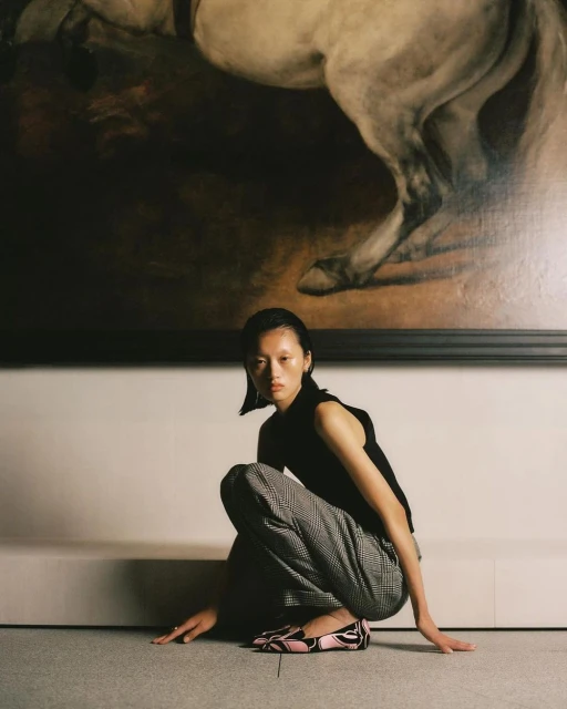 Fashion shot of an Asian woman in black top and plaid pants, squatting indoors in front of a painting of a white horse.