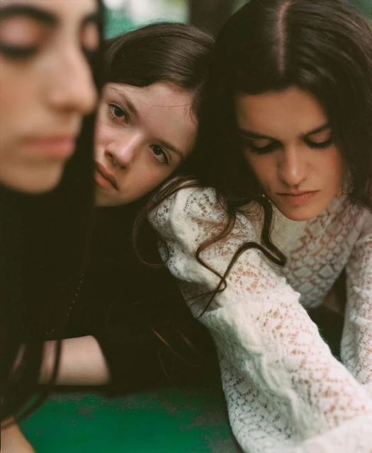 Three young women in a close indoor portrait. One looks at the camera, wearing a patterned lace top. The other two have dark hair.