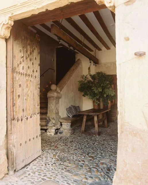 Open wooden door leads into stone-paved courtyard with rustic wooden table, stairs, and plant. Architectural interior detail.