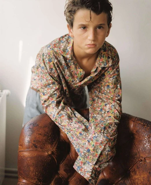 Portrait of a young boy in a floral button-down shirt leaning on a leather chair against a white wall with natural lighting.