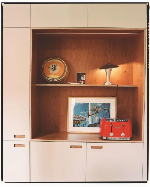 Retro interior of white cabinets with an orange clock, table lamp, framed photo, and red toaster against a warm wooden backdrop.