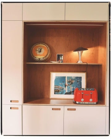Retro interior of white cabinets with an orange clock, table lamp, framed photo, and red toaster against a warm wooden backdrop.