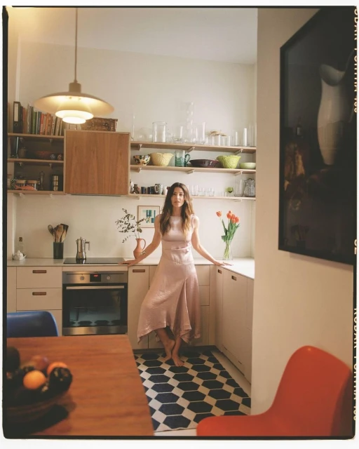 A woman in a dress stands barefoot in a bright kitchen with white walls, open shelves with dishware, and a hexagonal patterned floor mat.