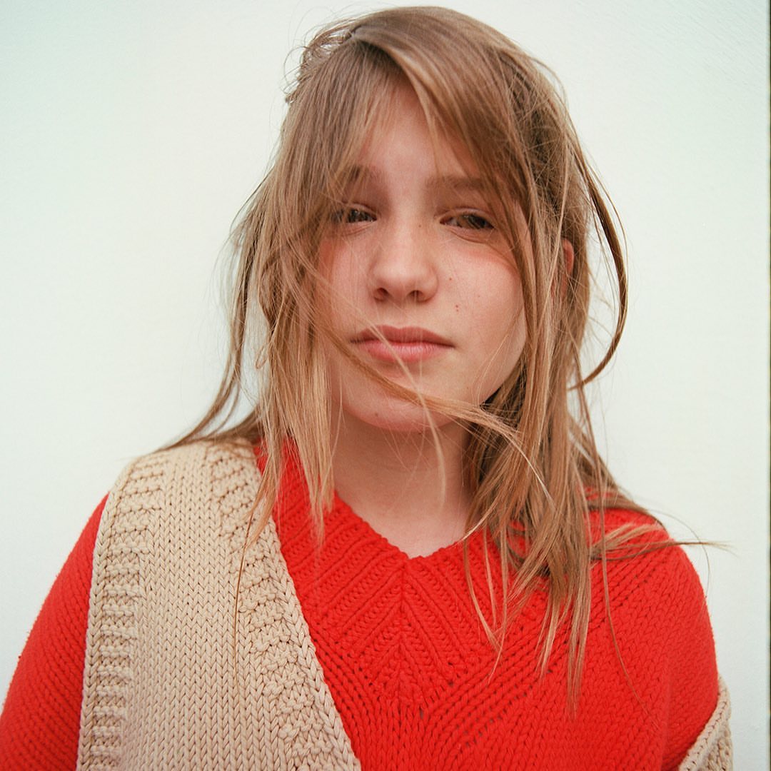 Close-up portrait of a young person with fair hair in a red knitted sweater and beige knit scarf posing against a white background.