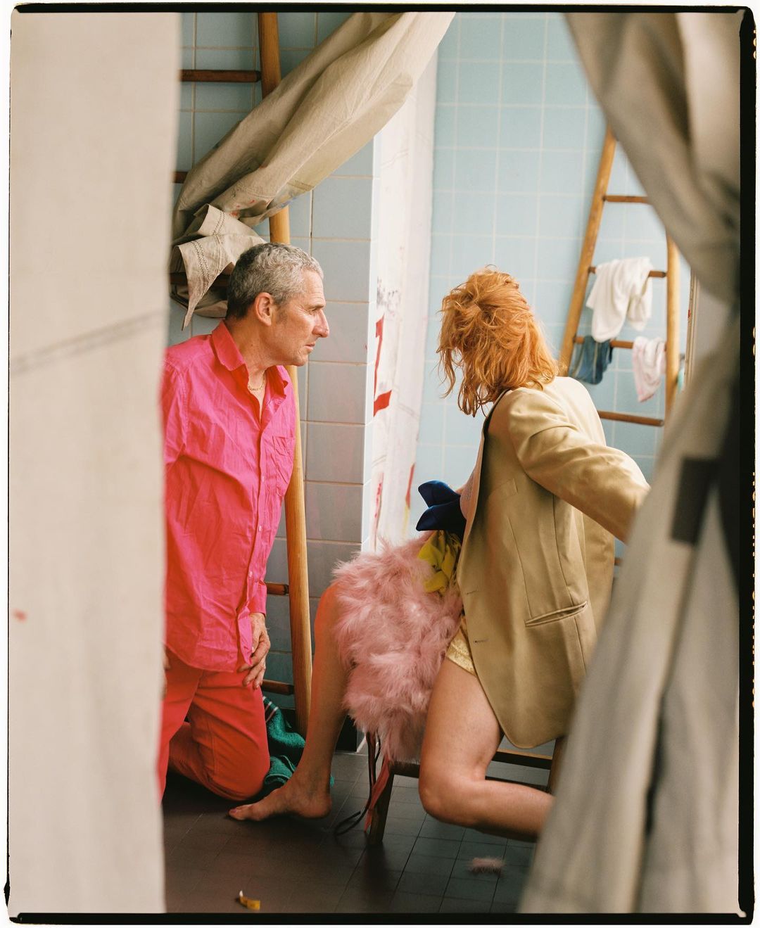 Indoor scene featuring a male in a pink pajama set looking at a red-haired woman in a blazer kneeling on a chair with a feathered prop.