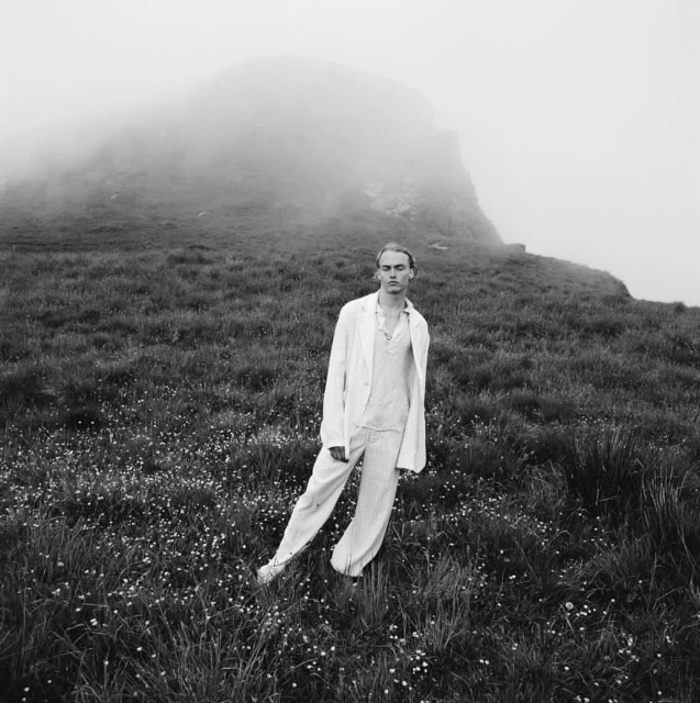A solitary figure stands in a grassy field near a mountain under a misty sky, dressed in a light-colored suit with a relaxed, open stance.