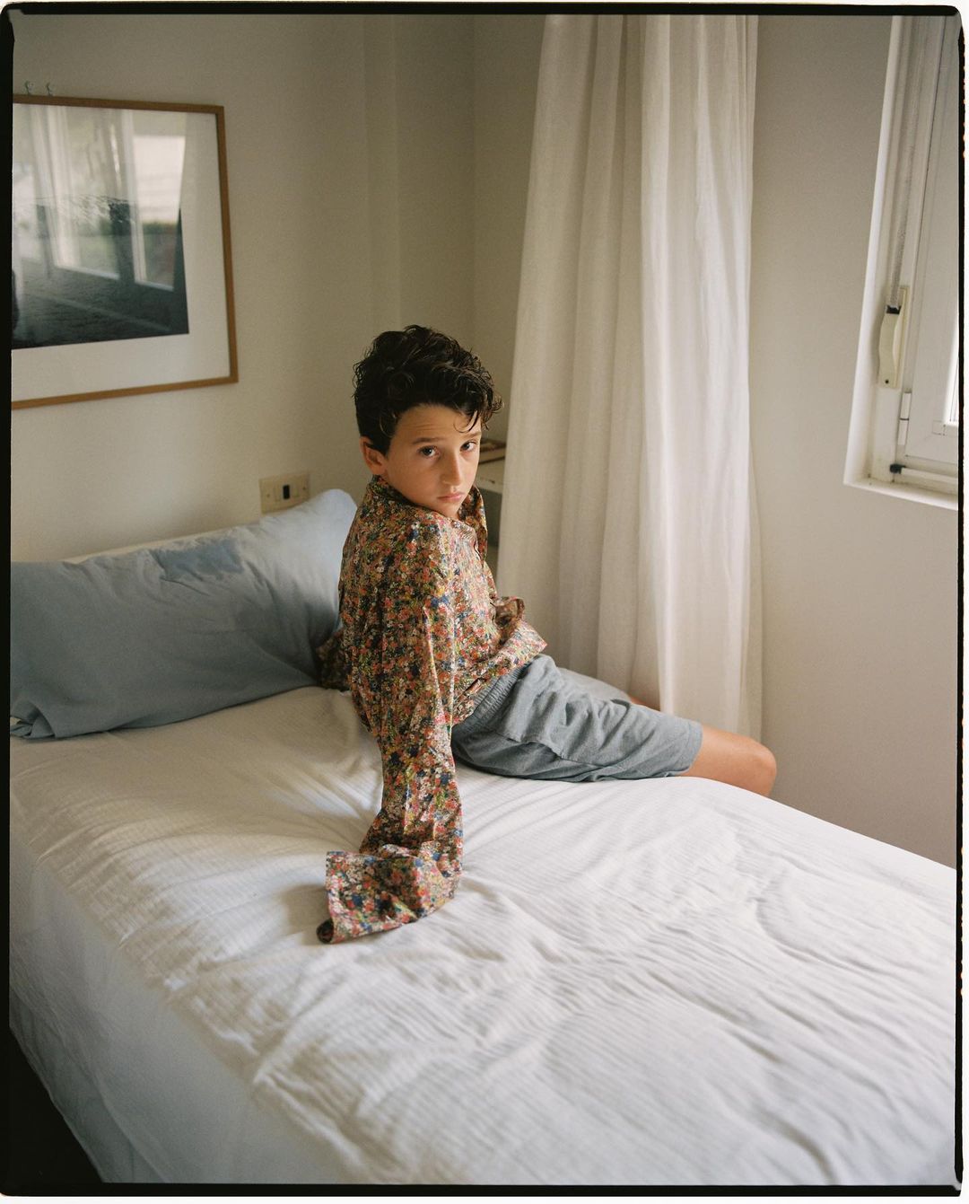 Boy sits on a white bed in a room with a framed picture, looking at the camera with a neutral expression. Natural light.