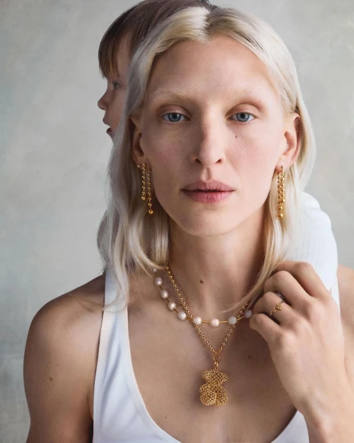 Close-up of a woman with blonde hair wearing gold jewelry including earrings and a teddy bear pendant necklace, with child behind her.