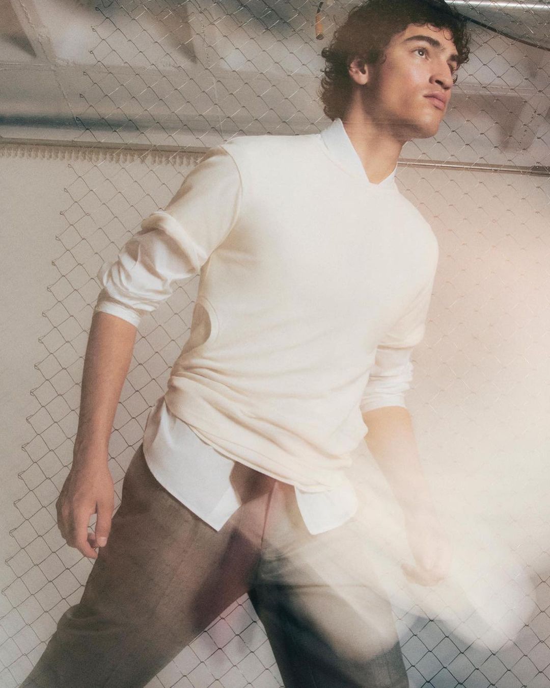 Adult male posing indoors, wearing white long-sleeve shirt and brown pants, lit by diffused light, set against a mesh background.