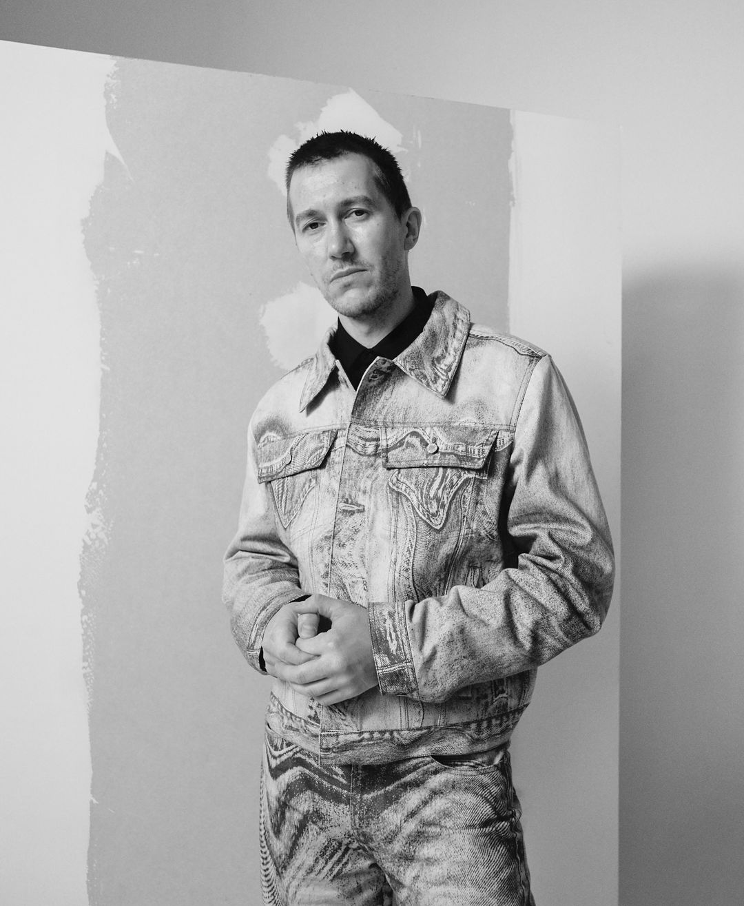 Black and white portrait of a man wearing a patterned denim jacket and pants, standing in front of a corner with light and shadow.