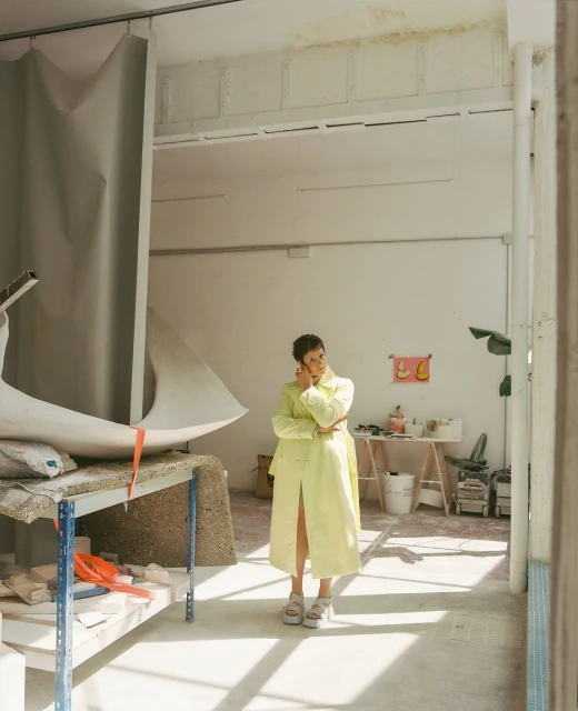 Woman in a chartreuse trench coat standing indoors in an artist's studio near abstract sculpture on a sunny day with white walls.