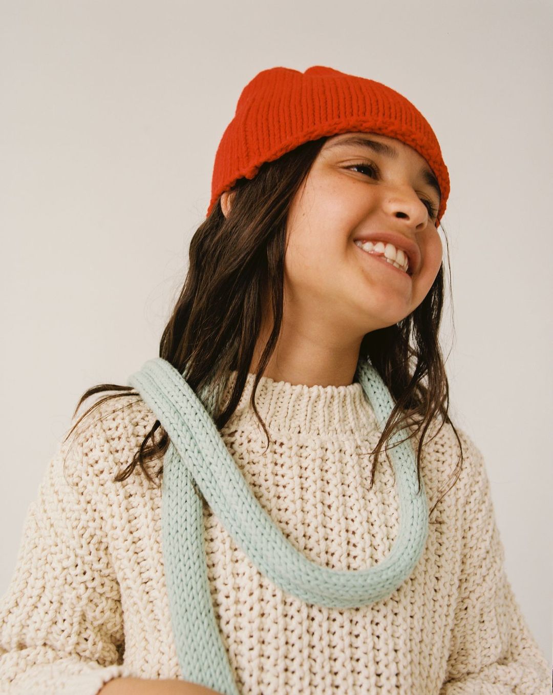 Smiling child portrait: A young girl with a red knit beanie, light sweater, and blue scarf smiles against a neutral studio backdrop.