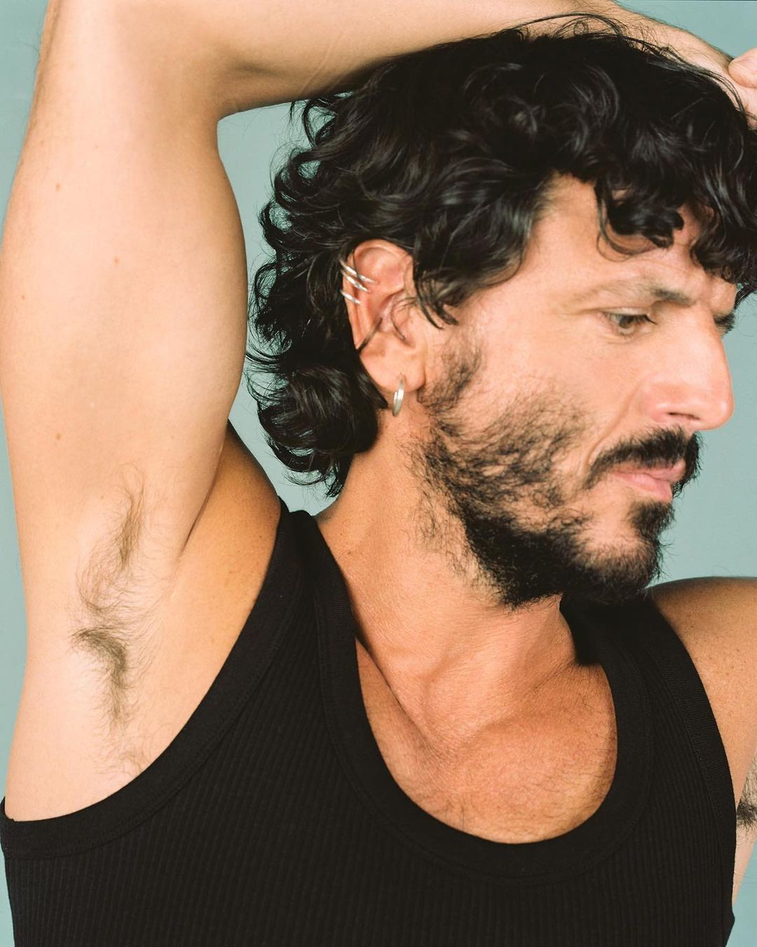 Close-up portrait of a man in a black tank top with dark curly hair and a beard, posing with his arm raised against a teal background.
