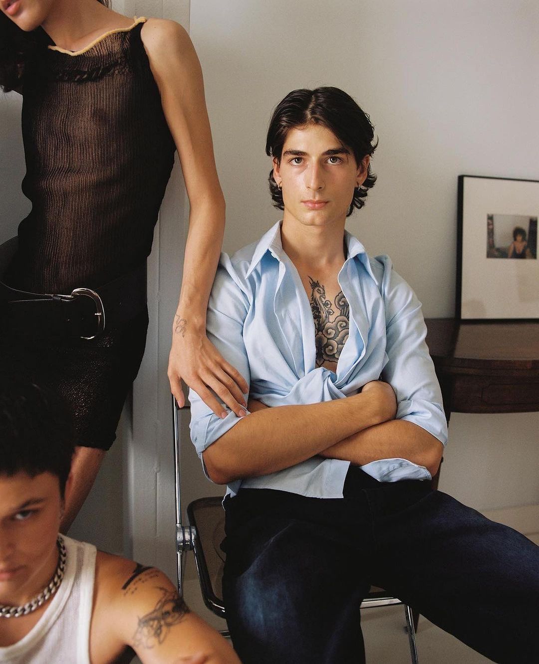 Three young adults are posing in an indoor setting. One person is seated and looking at the camera, while two are standing and leaning. 