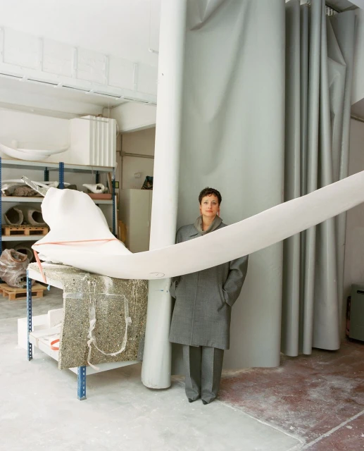 Woman in grey coat stands in an art studio with a large white abstract sculpture and white curtain. Indoor setting, neutral color palette.